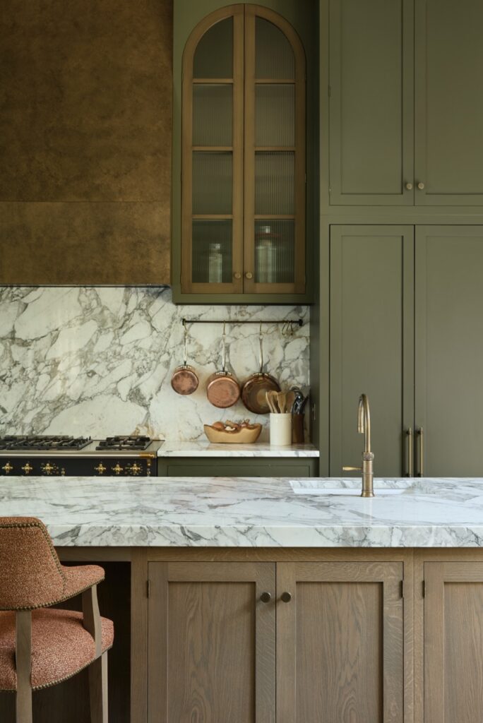 A modern kitchen with green cabinets, a marble backsplash and countertop, brass fixtures, copper pans hanging on the wall, and a wooden island with a brown upholstered chair. David Pearson Travel
