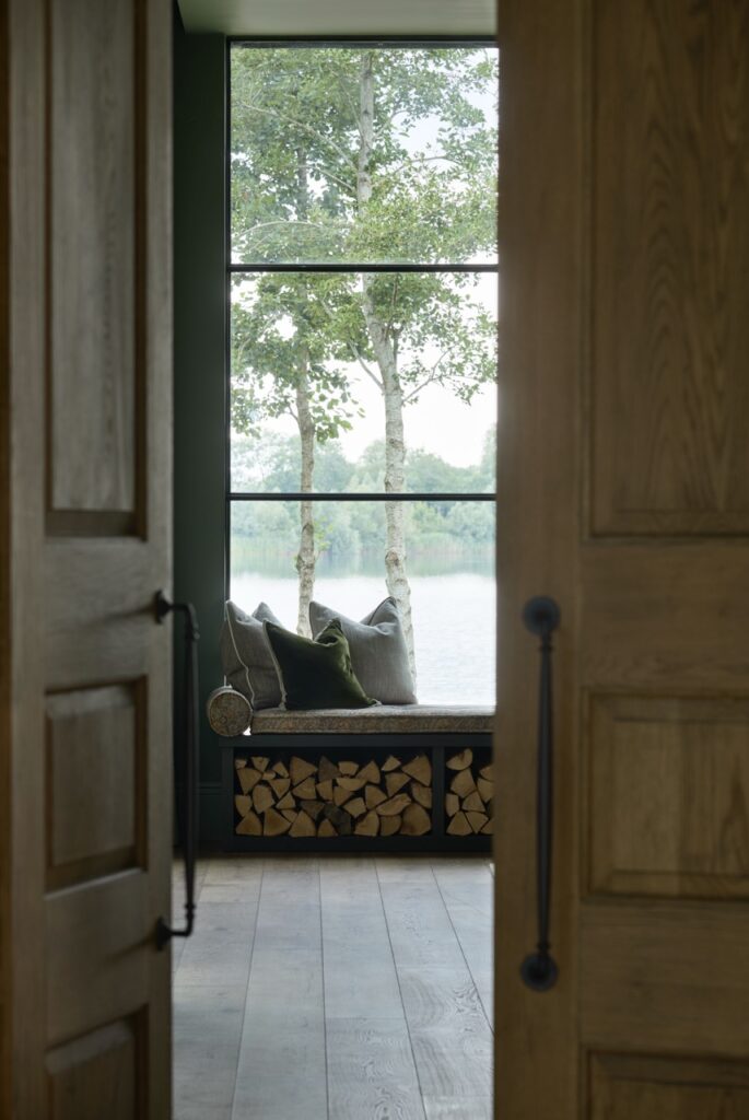 A cozy window seat with cushions overlooks trees and a lake, framed by large windows. Firewood is neatly stacked below the seat, and wooden double doors open into the room. David Pearson Travel