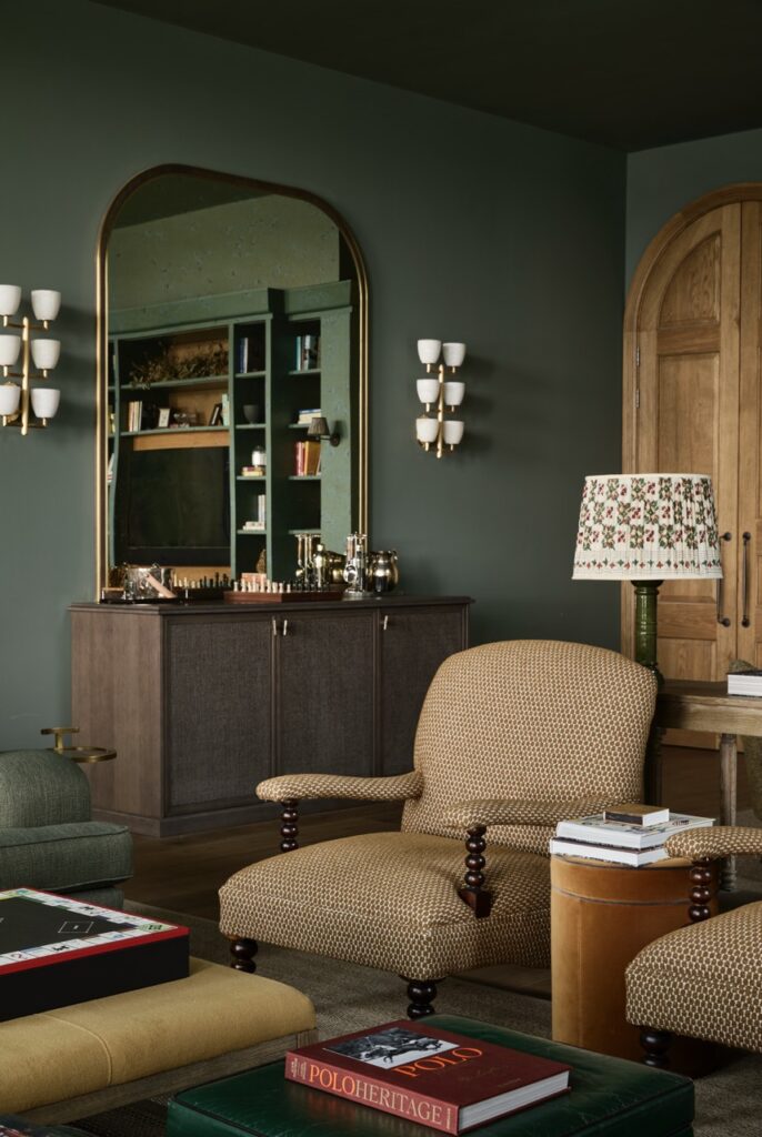 A cozy living room with dark green walls, a large arched mirror above a sideboard, patterned armchairs, a floral lampshade, built-in shelves, and wall sconces. A chess set and books are visible on the furniture. David Pearson Travel