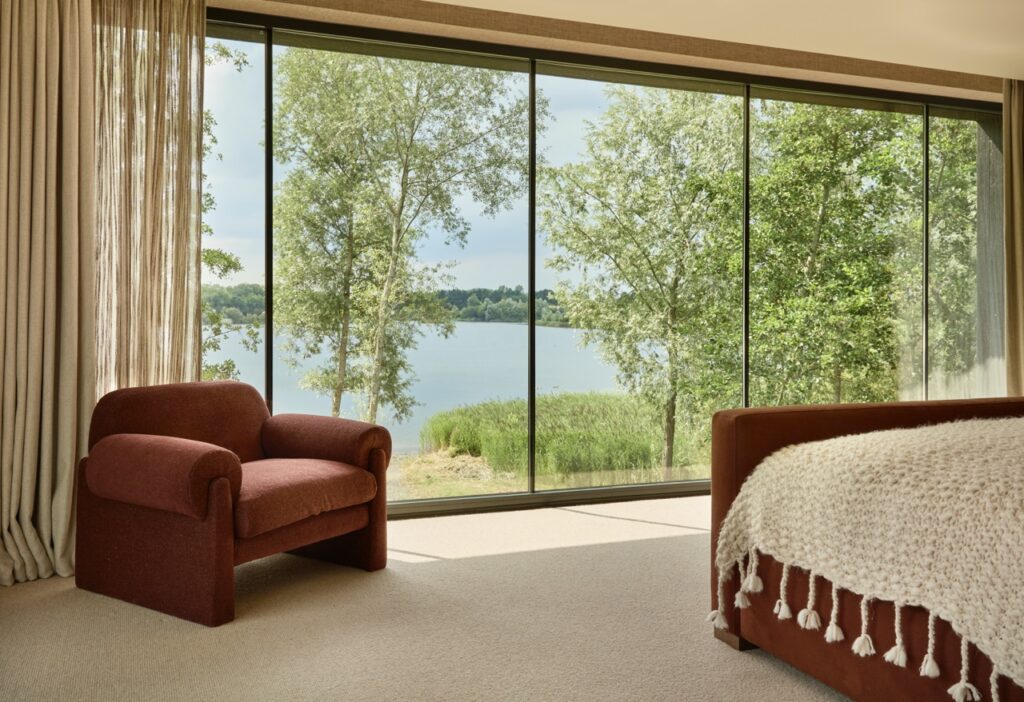 A cozy bedroom with a red armchair and a bed covered in a textured cream blanket, featuring floor-to-ceiling windows overlooking a lake and green trees outside. David Pearson Travel