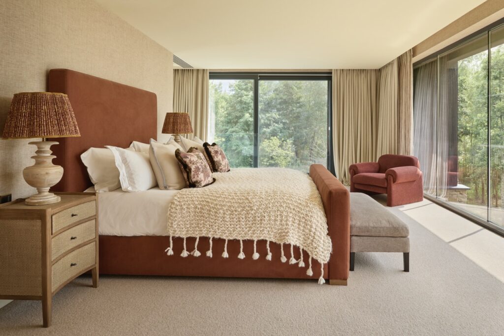 A spacious bedroom with a rust-colored upholstered bed, cream knitted blanket, patterned pillows, a wooden nightstand with a lamp, an upholstered bench, an armchair, large windows, and beige curtains overlooking greenery. David Pearson Travel