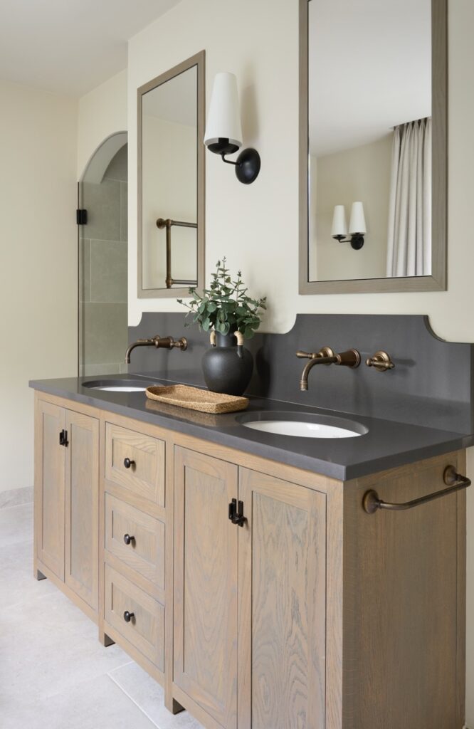 A modern bathroom with a double wood vanity, black countertop, two rectangular mirrors, wall-mounted faucets, a black vase with greenery, a woven tray, and light neutral walls with a towel bar on the side. David Pearson Travel