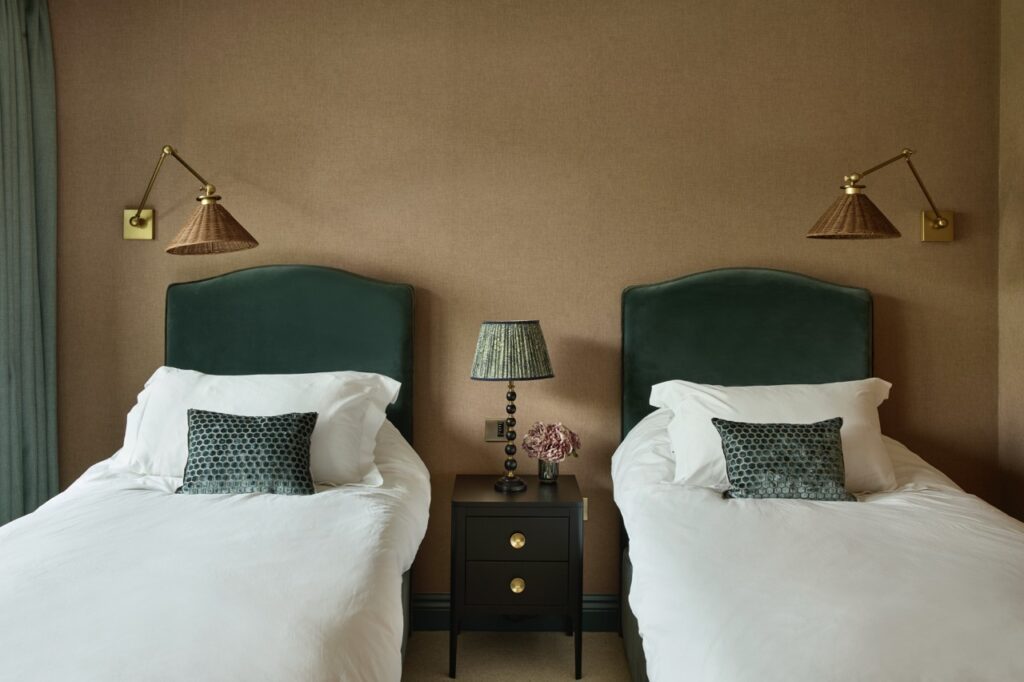 Two single beds with green velvet headboards and white bedding flank a small black nightstand with gold knobs, a lamp, and a vase of pink flowers. Woven wall lamps hang above each bed against a brown wall. David Pearson Travel