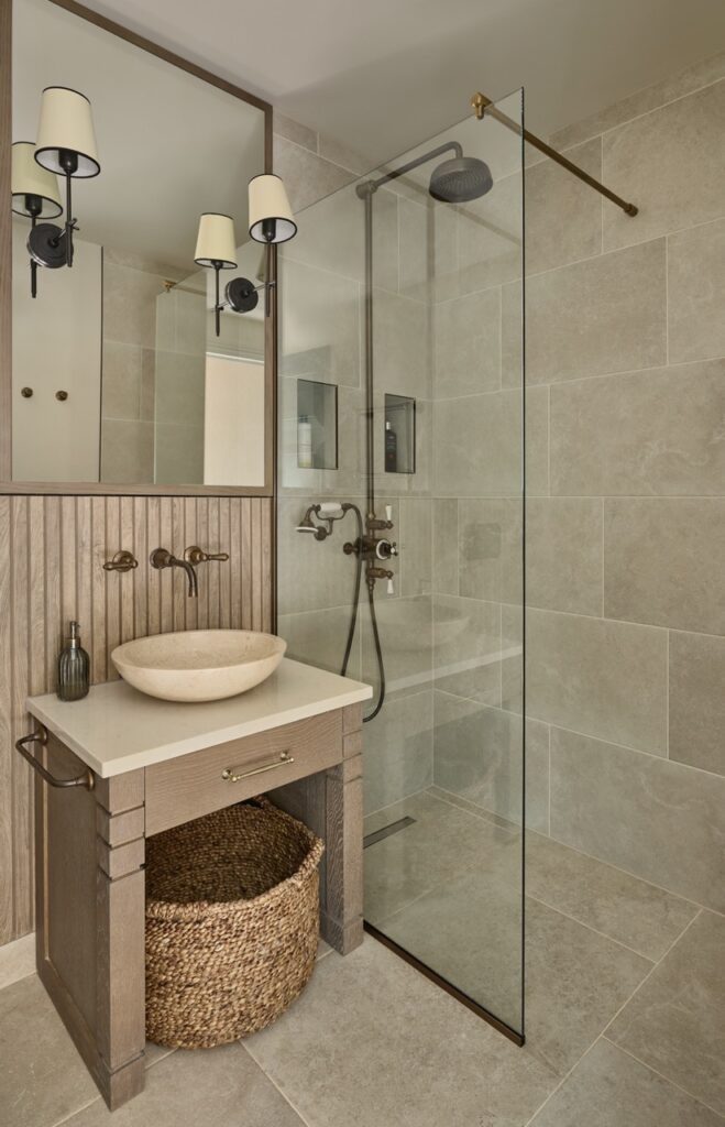 Modern bathroom with a stone sink on a wooden vanity, a wicker basket underneath, wall-mounted faucets, large mirror, walk-in glass shower with rainfall head, and beige tiled walls and floor. David Pearson Travel