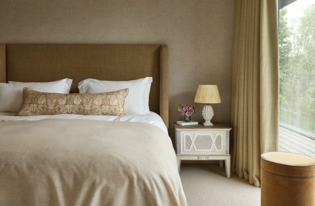 A cozy bedroom with a tan upholstered headboard, neutral bedding, a long decorative pillow, a white and wood nightstand holding a lamp and flowers, gold curtains, and a large window with a view of trees. David Pearson Travel