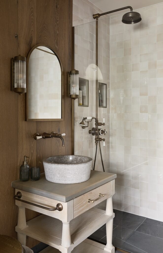 A modern bathroom with a stone vessel sink on a light wooden vanity, wall-mounted faucet, arched mirror, two wall sconces, and a walk-in shower with square white tiles and a dark rain showerhead. David Pearson Travel
