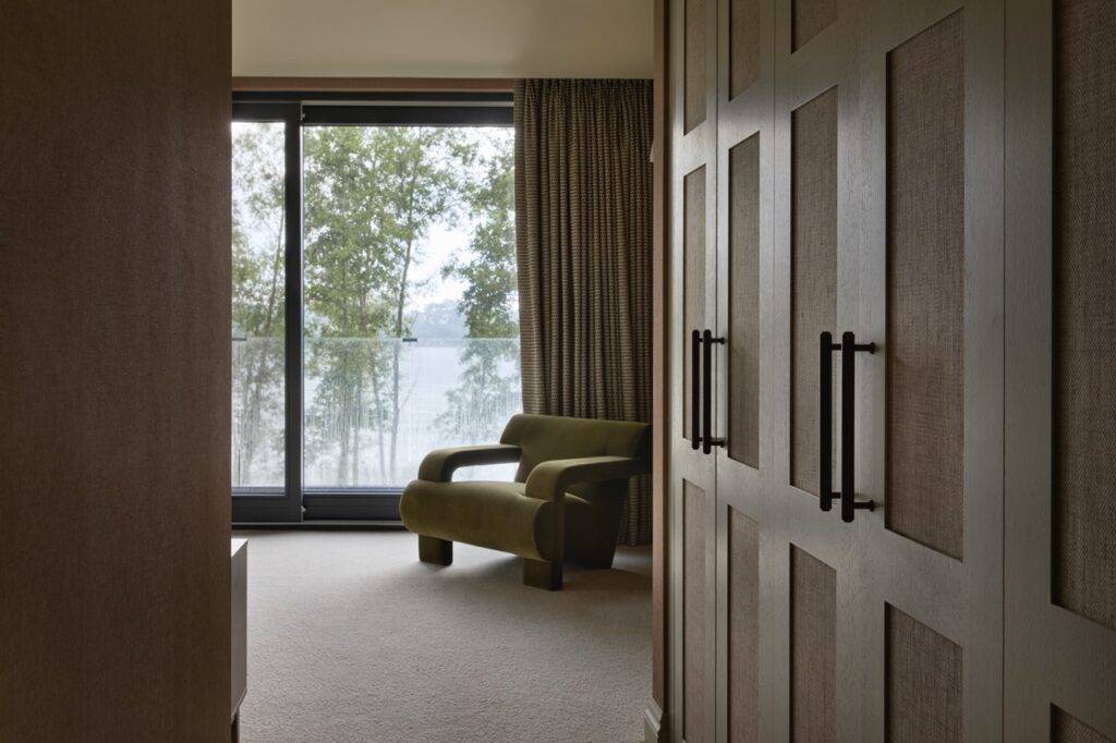 A modern bedroom with large built-in wooden closets, a green armchair near a floor-to-ceiling window with sheer curtains, and a view of trees and water outside. David Pearson Travel