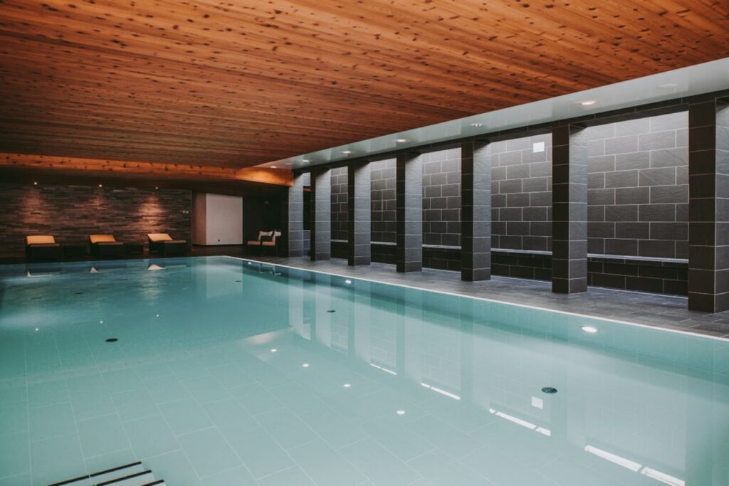 Indoor swimming pool with clear water, dark tiled walls, wooden ceiling, and lounge chairs along one side. The space is softly lit with recessed lighting. David Pearson Travel