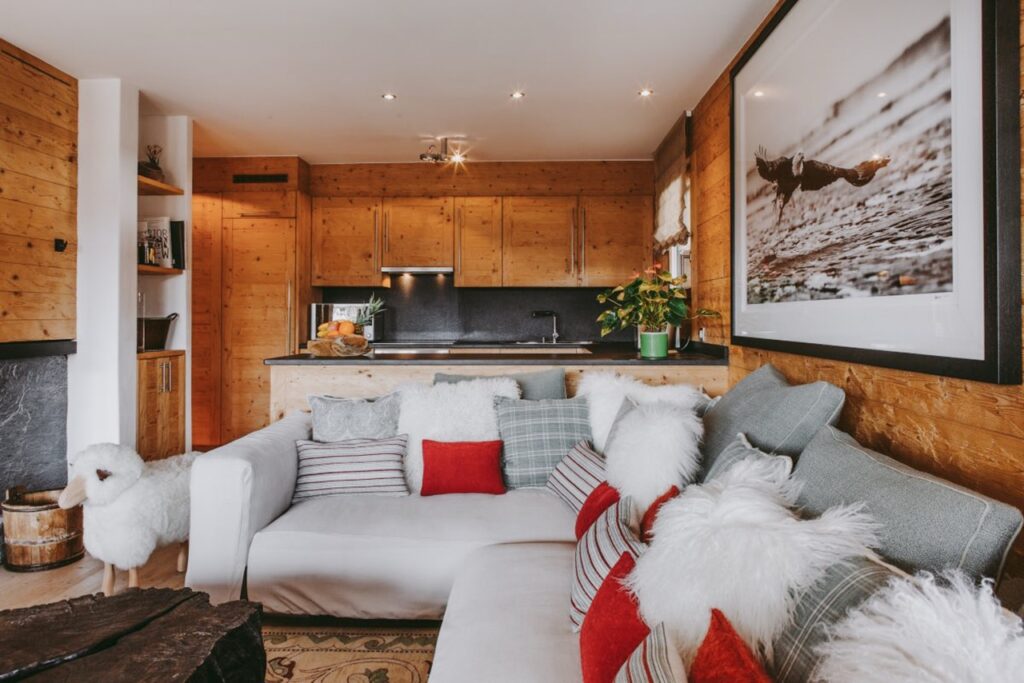 A cozy living room with a white couch decorated with red and white pillows, fluffy throws, wood-paneled walls, a sheep-shaped stool, and a modern kitchen in the background. A large framed photo hangs on the wall. David Pearson Travel