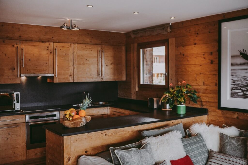 A cozy kitchen with wooden cabinets and walls, black countertops, a microwave, fruit bowl, and potted plant. There is a window, a framed photo on the wall, and a sofa with cushions and blankets in the foreground. David Pearson Travel