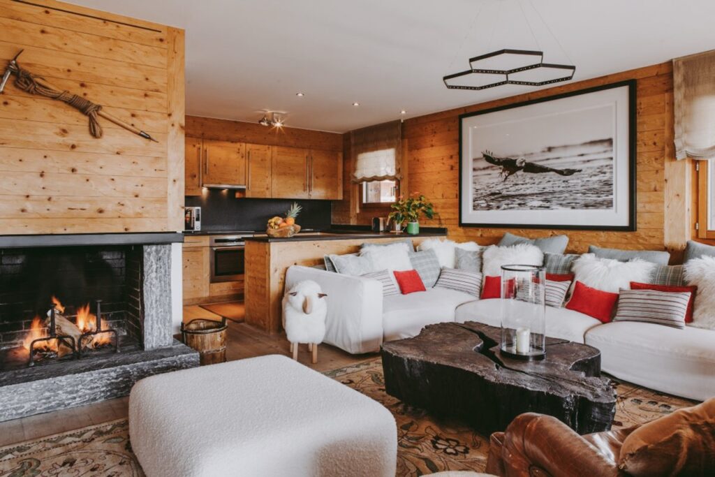 Cozy living room with wood-paneled walls, a lit stone fireplace, modern furniture, and a black coffee table. White sofas with red pillows face a large framed photo and an open kitchen with wood cabinets in the background. David Pearson Travel
