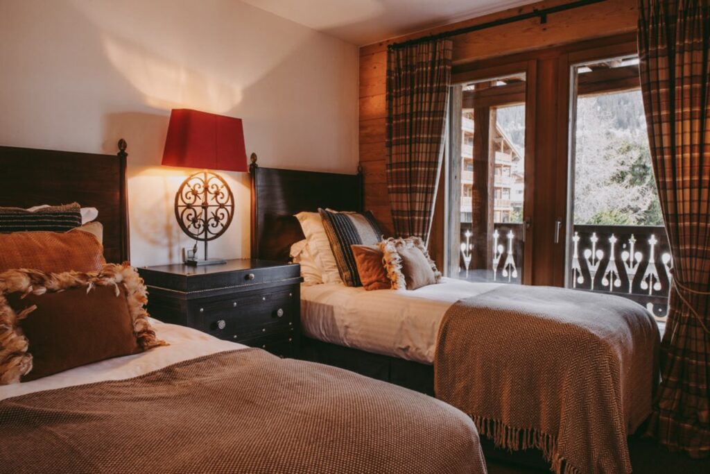Cozy bedroom with two single beds, brown and beige bedding, fluffy pillows, a dark nightstand, ornate lamp, and large windows with curtains overlooking a balcony and mountain scenery. David Pearson Travel