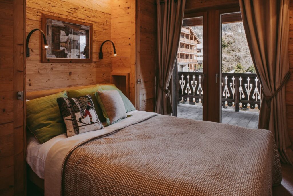 Cozy wooden bedroom with a double bed, green and beige pillows, reading lamps, and a picture on the wall. Large glass doors open to a balcony with mountain and chalet views outside. Curtains are pulled to the sides. David Pearson Travel