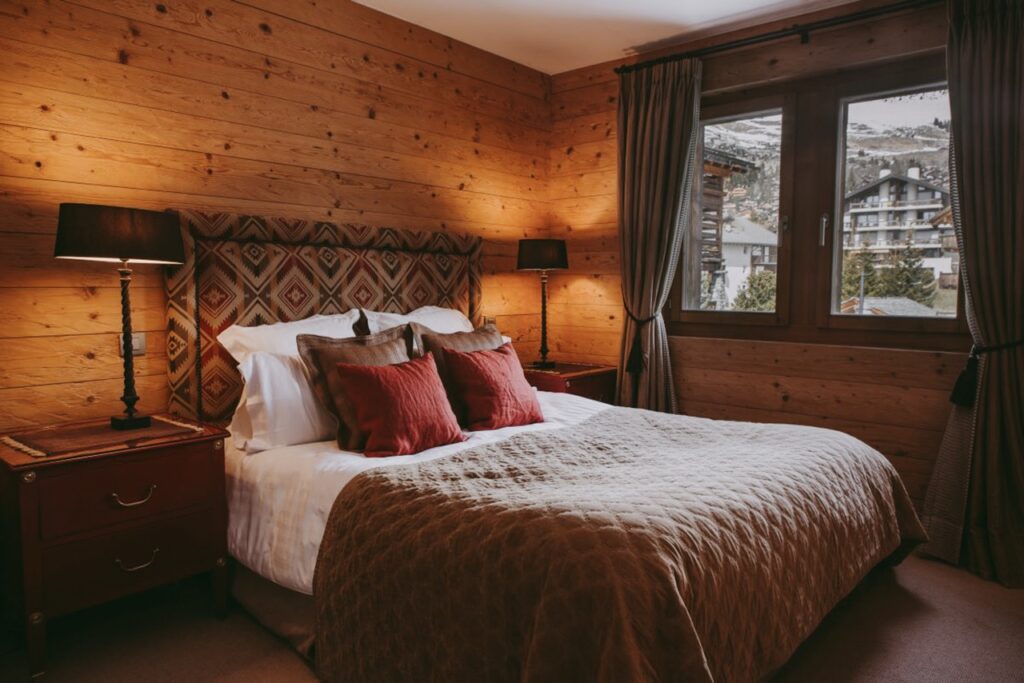 A cozy bedroom with wooden walls, a large bed with a patterned headboard, white linens, brown quilt, and red pillows. Two bedside tables with lamps are beside the bed, and a window shows a snowy mountain scene outside. David Pearson Travel