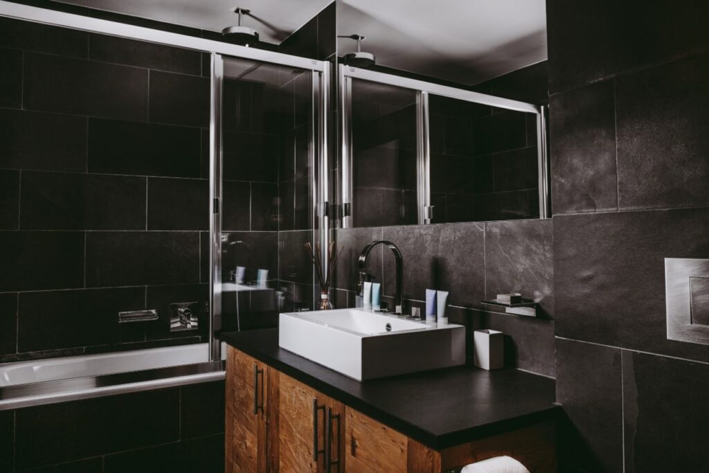 Modern bathroom with black tiled walls, a wooden vanity with a square white sink, chrome faucet, and toiletries. Large mirrors and a glass-enclosed bathtub are also visible, creating a sleek, contemporary look. David Pearson Travel