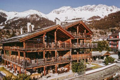 Large wooden chalet with balconies, set against snow-capped mountains and pine trees under a clear sky, surrounded by other alpine buildings. David Pearson Travel