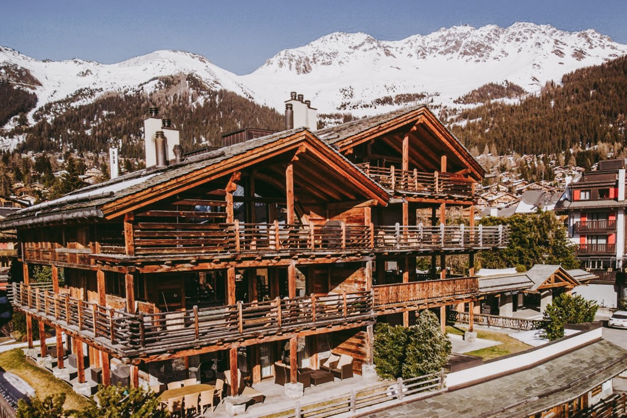 Large wooden chalet with balconies, set against snow-capped mountains and pine trees under a clear sky, surrounded by other alpine buildings. David Pearson Travel