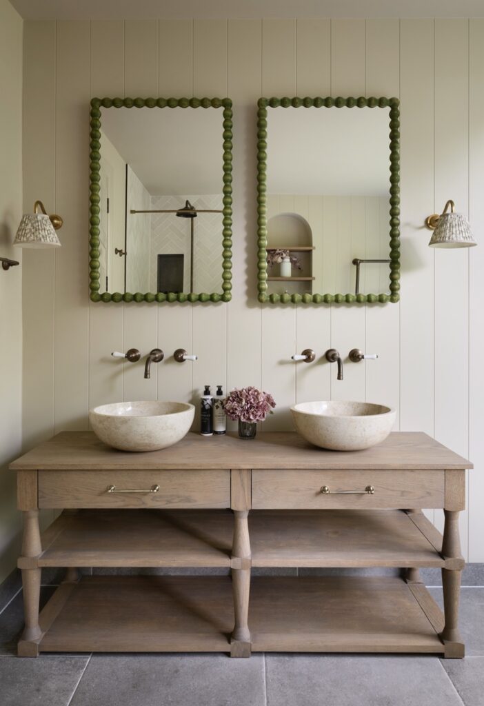 A bathroom with a double wooden vanity, two round vessel sinks, two green-framed mirrors, wall-mounted faucets, sconces, and a vase with pink flowers. Neutral colors and a minimalist design. David Pearson Travel