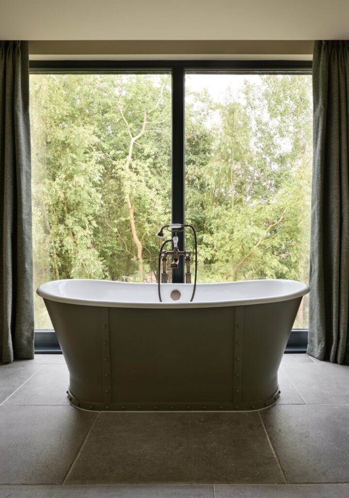 A freestanding bathtub sits on gray tiles in front of a large window with lush green trees outside, framed by dark curtains on each side, creating a tranquil bathroom setting. David Pearson Travel