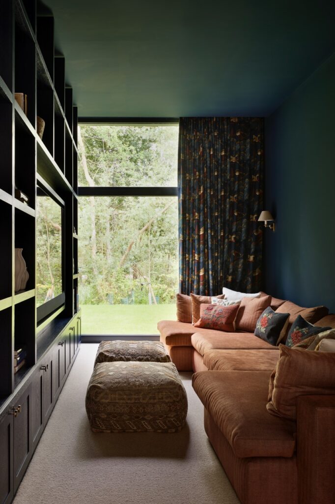 A cozy living room with a large sectional sofa in earthy tones, patterned cushions, two ottomans, a dark built-in bookcase, and a big window with a forest view, filtered by floral curtains. David Pearson Travel