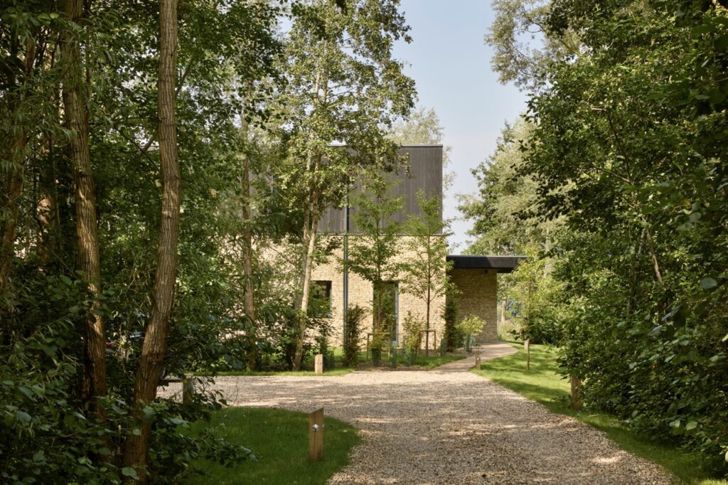 A modern house with stone walls and a dark roof is surrounded by tall trees and lush greenery, with a gravel path leading up to the entrance on a sunny day. David Pearson Travel