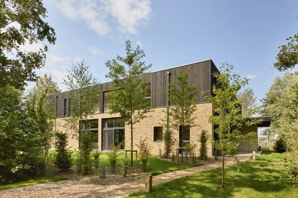 A modern two-story house with a brick lower level and dark wood upper level, surrounded by green grass, young trees, and shrubs under a partly cloudy sky. David Pearson Travel