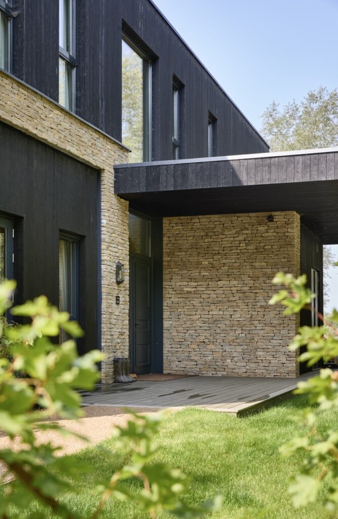 Modern house exterior with dark wood siding and light stone walls, featuring large windows, covered entryway, and green lawn with leafy plants in the foreground. David Pearson Travel