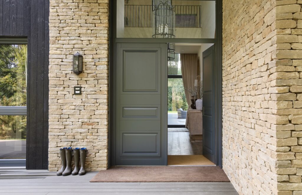 A modern house entrance with a partially open dark gray door set in a wall of light-colored stone. Three pairs of rubber boots are lined up on the left by the door, and a doormat lies at the threshold. David Pearson Travel