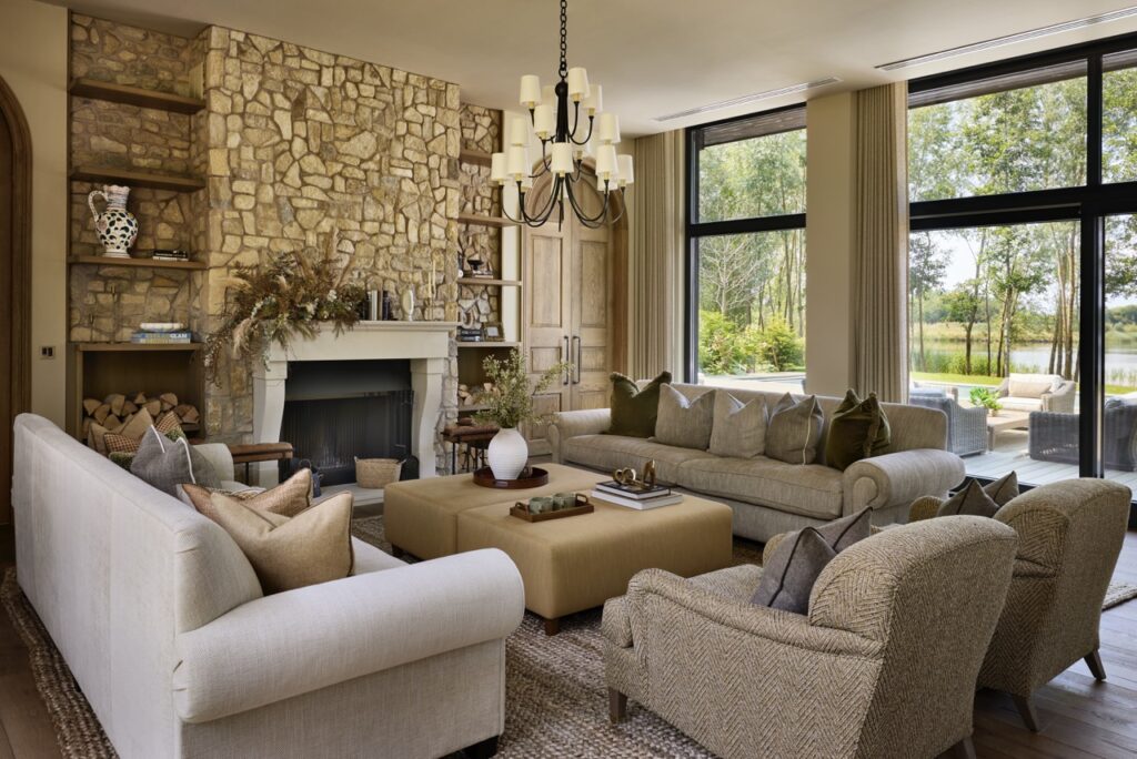 A cozy living room with two sofas and two armchairs around a tan ottoman, a stone fireplace, built-in shelves, large windows with a view of trees and water, and neutral décor with earthy accents. David Pearson Travel