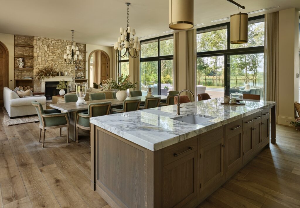 Open-concept kitchen and living area with a marble island, wooden floors, a dining table with green chairs, stone fireplace, and large windows overlooking a green outdoor space. Neutral tones and natural light fill the room. David Pearson Travel