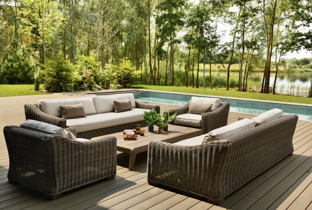 Four wicker sofas with white cushions are arranged around a wooden coffee table on a deck beside a pool, surrounded by green trees and overlooking a tranquil lake. David Pearson Travel