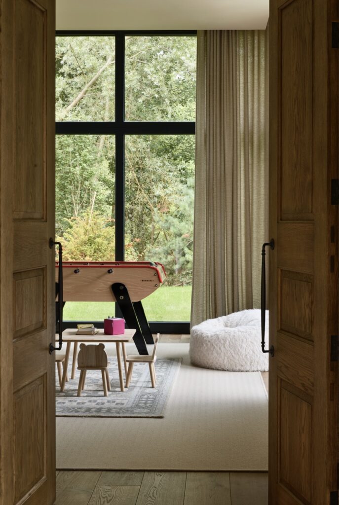 A cozy room with tall windows, a small air hockey table, a low table with chairs, a pink bucket, a plush bean bag chair, beige curtains, and a view of greenery outside. David Pearson Travel