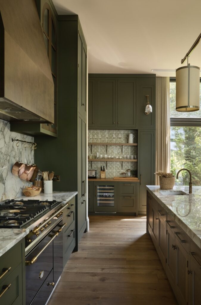 A modern kitchen with olive green cabinets, marble countertops, a gas stove with brass accents, a farmhouse sink, wooden floors, and natural light coming through large windows. Copper cookware hangs on the backsplash. David Pearson Travel