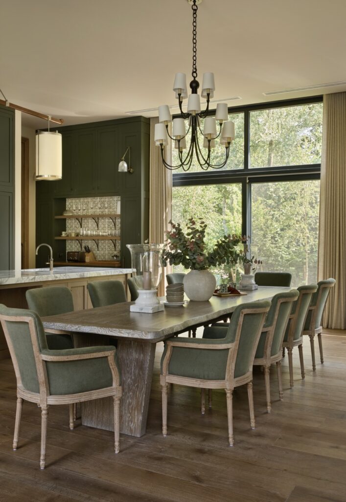 A dining room with a large wooden table, eight green cushioned chairs, a modern chandelier, and vases with flowers. Large windows with beige curtains let in natural light and show trees outside. The kitchen is visible in the background. David Pearson Travel