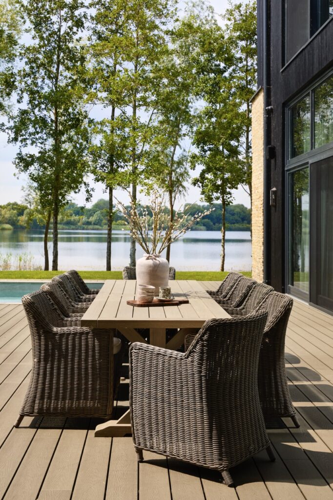 Outdoor patio with wicker chairs around a wooden table, a vase with branches as centerpiece, and a scenic lake and trees in the background on a sunny day. David Pearson Travel