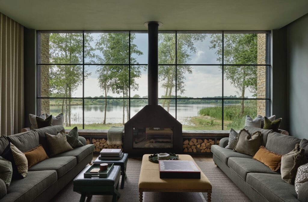 A modern living room with two gray sofas, a central fireplace, large windows revealing a lake and trees outside, and neatly stacked firewood under the windows. Pillows and books decorate the space. David Pearson Travel