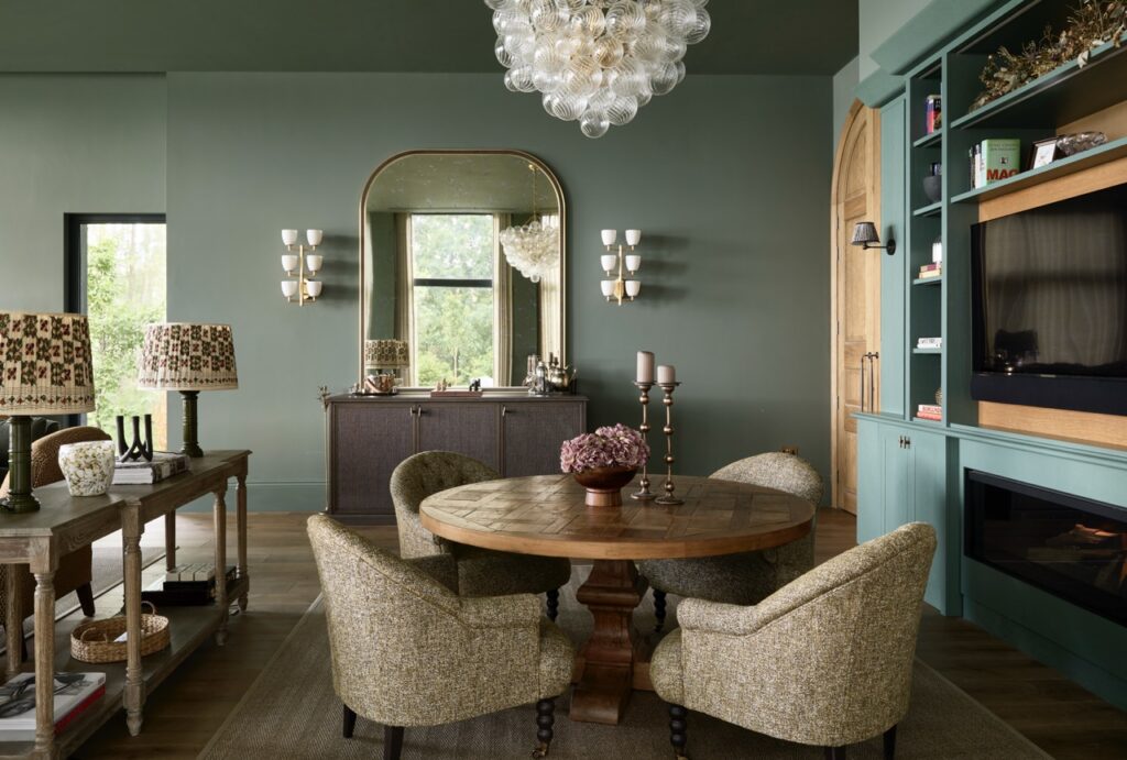 A stylish dining room with green walls, a round wooden table surrounded by four textured chairs, a large mirror, sideboard, built-in shelves, chandelier, and decorative lamps, all creating a cozy, elegant atmosphere. David Pearson Travel
