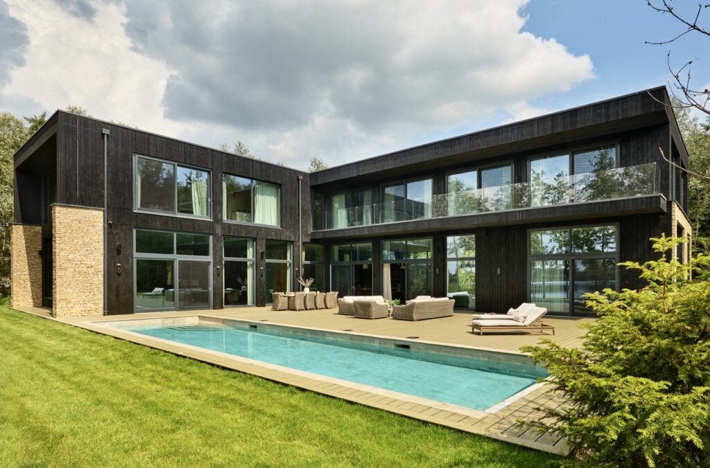 Modern two-story house with dark wood exterior, large glass windows, and a stone accent wall, featuring a backyard patio with outdoor furniture and a rectangular swimming pool, surrounded by a green lawn. David Pearson Travel