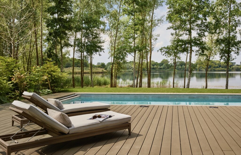 Two cushioned lounge chairs sit on a wooden deck beside a swimming pool, overlooking a scenic lake surrounded by trees and greenery under a bright, partly cloudy sky. David Pearson Travel