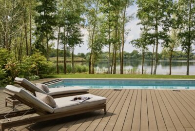 Two cushioned lounge chairs sit on a wooden deck beside a swimming pool, overlooking a scenic lake surrounded by trees and greenery under a bright, partly cloudy sky. David Pearson Travel