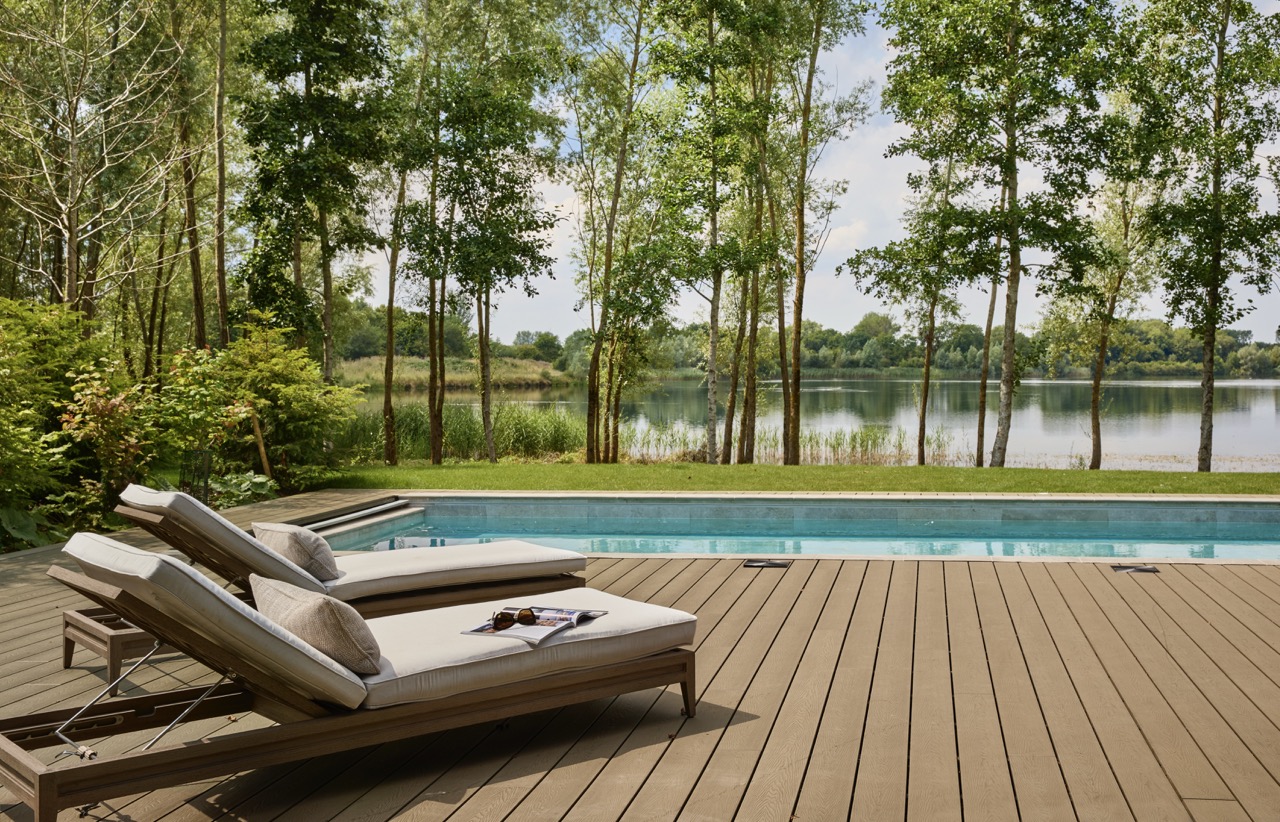 Two cushioned lounge chairs sit on a wooden deck beside a swimming pool, overlooking a scenic lake surrounded by trees and greenery under a bright, partly cloudy sky. David Pearson Travel