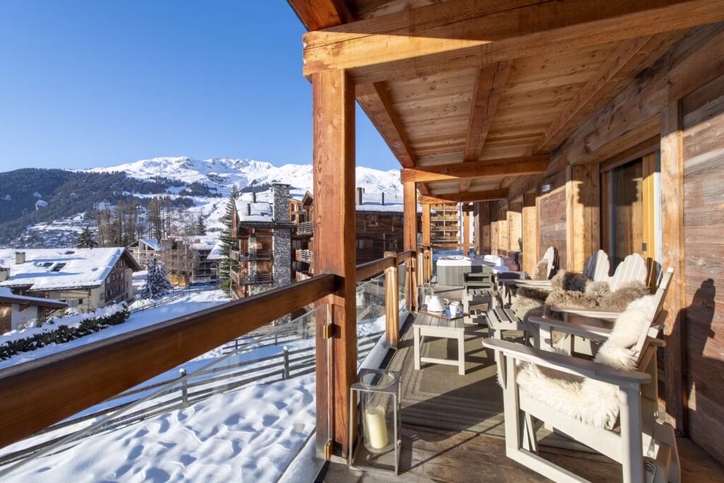 A wooden chalet balcony with cozy chairs and blankets overlooks snow-covered rooftops and mountains under a clear blue sky. Lanterns and rustic decor add warmth to the winter scene. David Pearson Travel