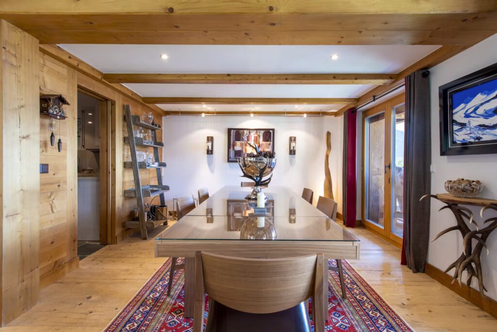 A modern dining room with wooden beams and floor, a glass table with six chairs, art on the walls, shelves with decor, and large windows letting in natural light. A patterned rug adds color beneath the table. David Pearson Travel