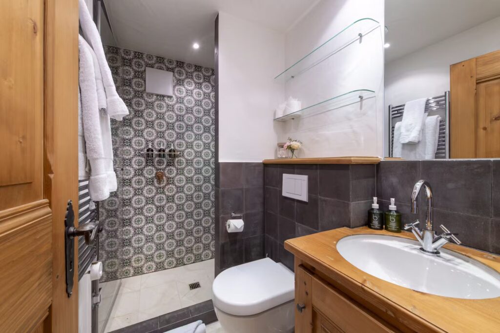A modern bathroom with a patterned tile walk-in shower, glass shelves, a toilet, a wooden vanity with a sink, and toiletries. Towels are hung on racks, and a small vase with flowers sits by the toilet. David Pearson Travel