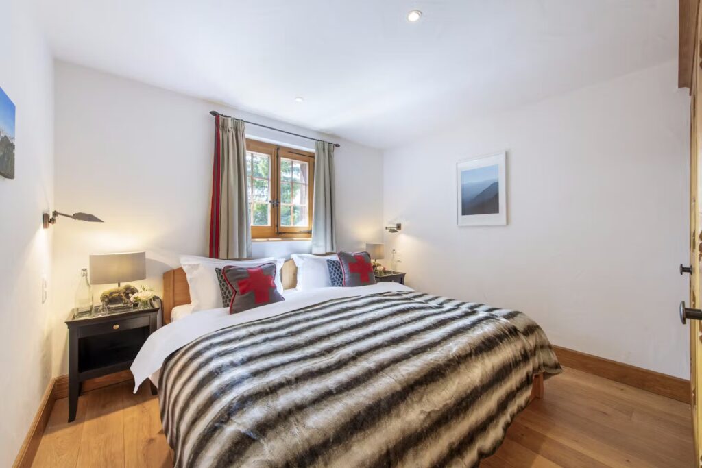 A cozy bedroom with a large bed covered by a striped fur blanket, two nightstands with lamps, pillows with red accents, a window with curtains, a wall-mounted photo, and wooden floors. David Pearson Travel