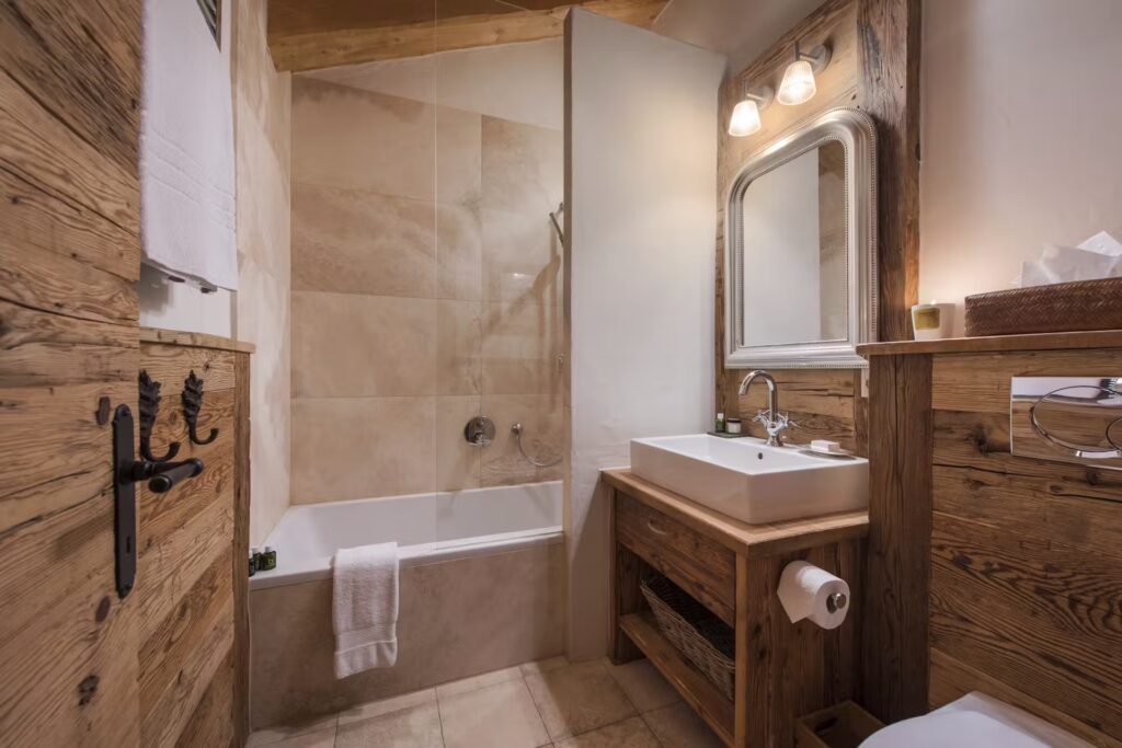 Rustic bathroom with wooden walls and fixtures, a white sink with a large mirror, a bathtub with glass screen, beige tiles, and soft lighting creating a cozy atmosphere. David Pearson Travel