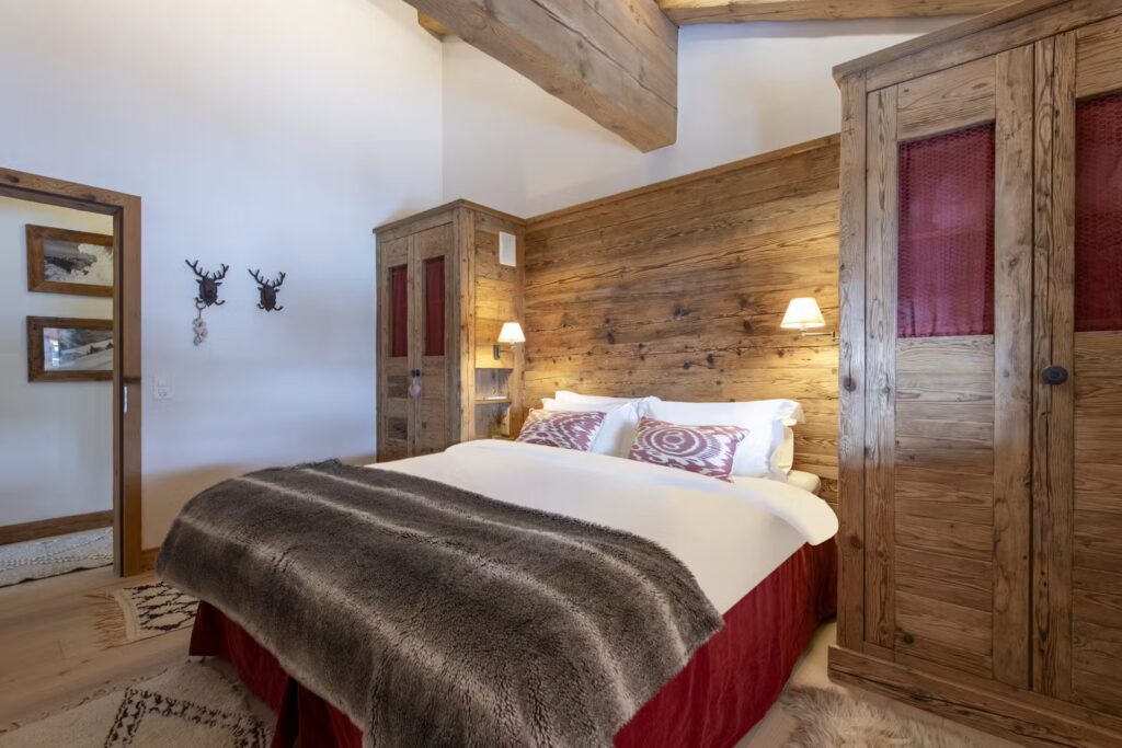 A cozy bedroom with rustic wooden walls, a large bed with patterned pillows and a faux fur throw, two wooden wardrobes, soft lighting, and decorative antler hooks on the wall. David Pearson Travel