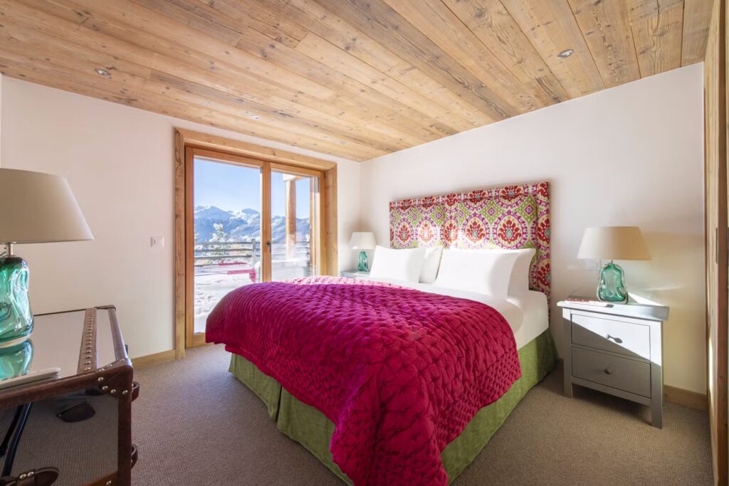 A cozy bedroom with a colorful patterned headboard, a large bed covered in a bright red quilt, two nightstands with lamps, a glass-topped desk, and glass doors opening to a mountain view with snow outside. David Pearson Travel
