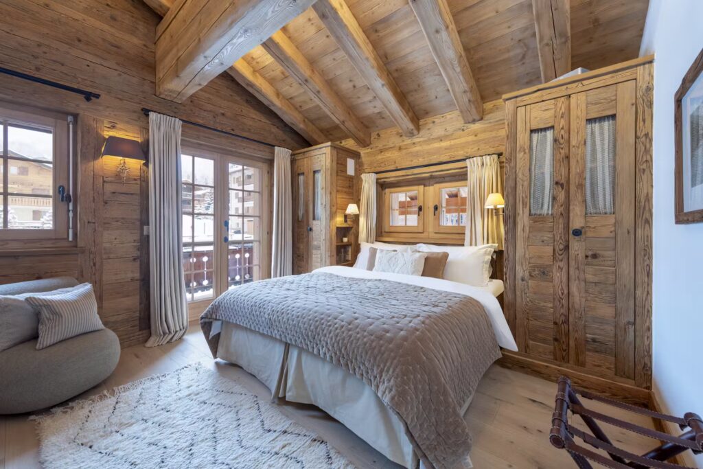 Cozy bedroom with rustic wooden walls and ceiling, a large bed with gray bedding, soft lighting, and windows letting in natural light. There’s a plush chair, a woven rug, and a view of snowy chalets outside. David Pearson Travel