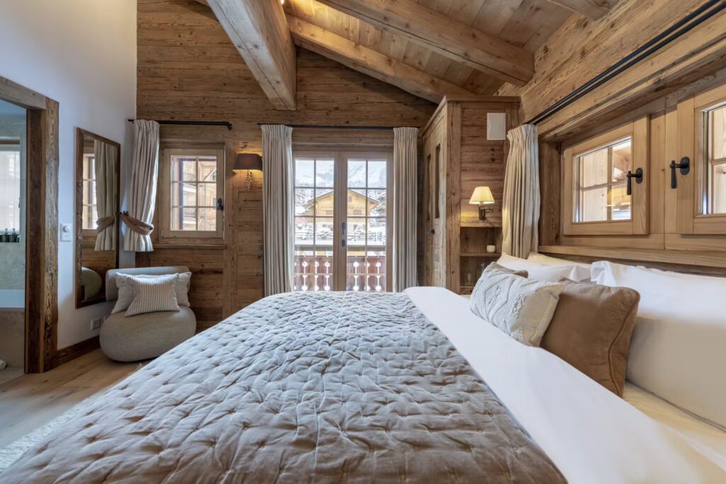 Cozy bedroom with wooden walls and ceiling, a large bed with beige and brown bedding, windows with curtains, a balcony door, and an armchair in the corner. Natural light brightens the rustic, inviting space. David Pearson Travel